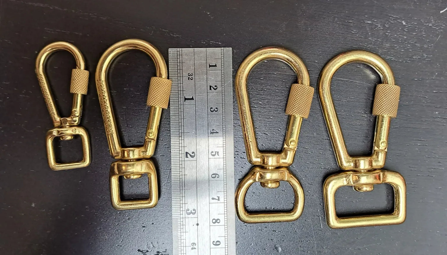 Four gold metal swivel hooks with a ruler for scale on a dark surface