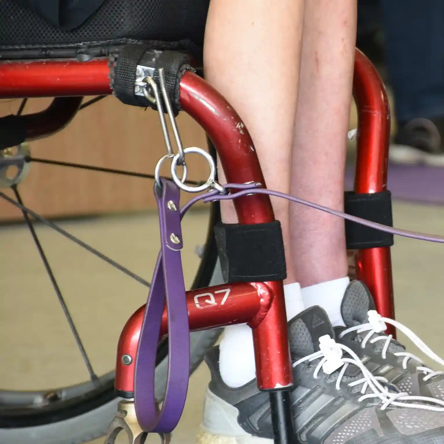 close up of a red manual wheelchair showing the Hook Attachment holding a purple leash, person's legs and gray shoes.